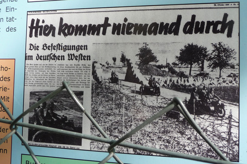 Hinter Maschendrahtzaun ist ein Zeitungsartikel zu sehen, der auf einer Tafel des Westwall-Wanderwegs Oberotterbach abgedruckt ist. Der Titel des Artikels lautet "Hier kommt niemand durch."