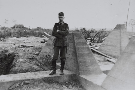 Ein junger Soldat steht beim Höckerlinienbau mit verschränkten Armen zwischen den Höckern. Im Hintergrund ist die Panzerkuppel eines Bunkers zu sehen.