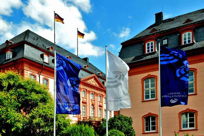 Im Hintergrund befindet sich das Gebäude des rheinland-pfälzischen Landtages. Im Vordergrund wehen weiße und blaue Fahnen. 