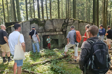Patrice Wijnands steht im lichten Nadelwald vor einem nur leicht gesprengten Bunker und referiert vor mehreren Personen über den Bunker und die historischen Zusammenhänge.